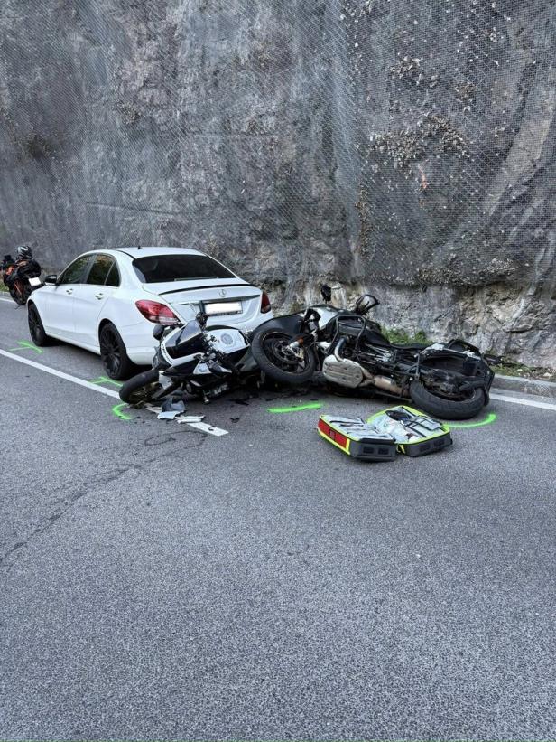 Ein weißes Auto und zwei gestürzte Motorräder liegen nach einem Unfall auf einer Straße vor einer Felswand.