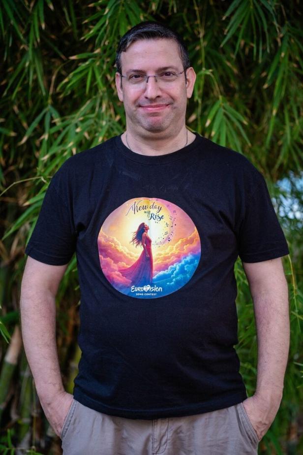 Ein Mann mit Brille trägt ein schwarzes T-Shirt mit farbigem Eurovision-Motiv und steht vor grünem Bambus.
