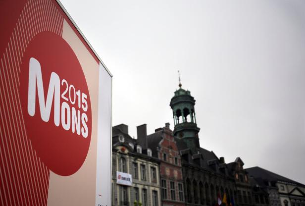 Ein Schild für „Mons 2015“ vor einer historischen Stadtansicht.