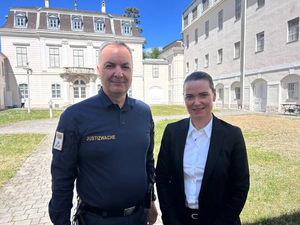 Zwei Personen, eine in Justizwache-Uniform, stehen vor einem hellen Gebäude.