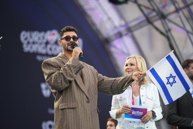 Ein Mann mit Mikrofon und Sonnenbrille steht neben einer Frau, die eine israelische Flagge hält, auf einer Bühne beim Eurovision Song Contest.