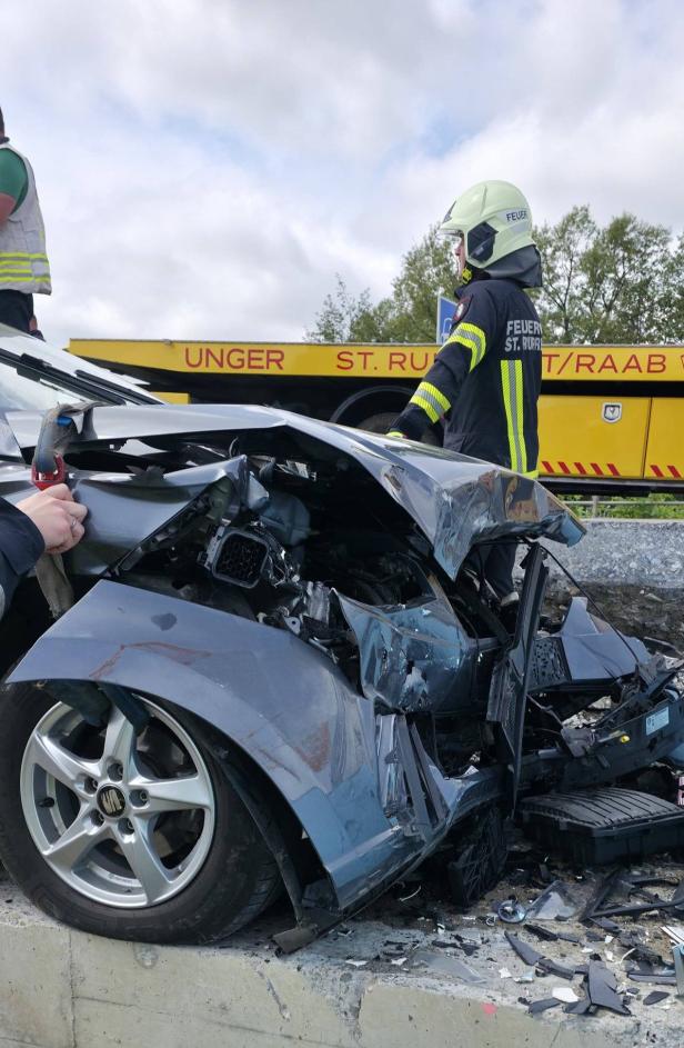 Stark beschädigtes Auto nach Unfall, Feuerwehrmann und Abschleppfahrzeug im Hintergrund.