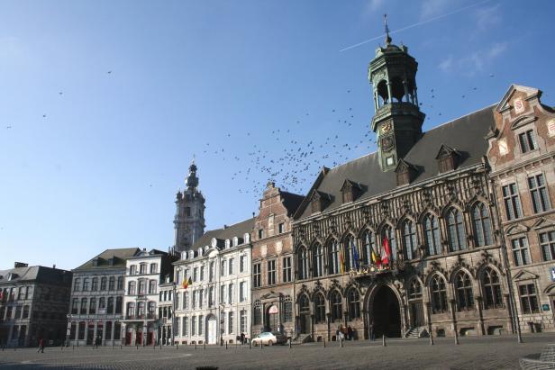 Der Grand Place in Mons, Belgien, mit historischer Architektur und einem Vogelschwarm.