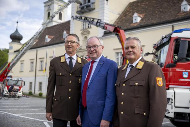 drei Männer, zwei in Uniform, einer in blauem Anzug stehen vor zwei Feuerwehrautos vor dem Stift Heiligenkreuz.