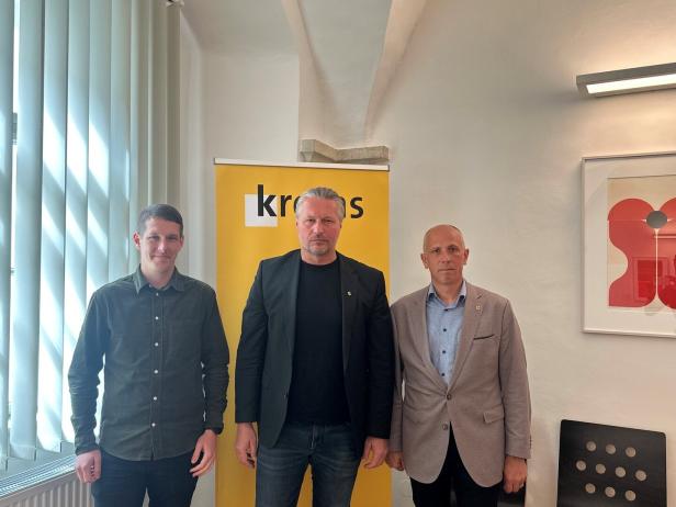 Drei Männer stehen nebeneinander vor einem gelben Aufsteller mit der Aufschrift „kro...“ in einem hellen Raum.