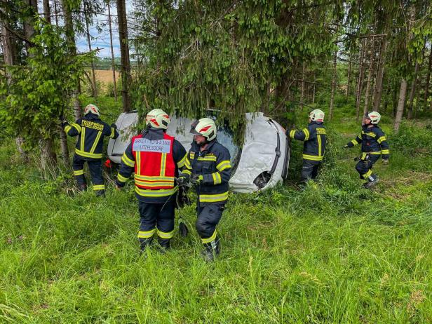 Mehrere Feuerwehrleute bergen ein auf der Seite liegendes Auto im Wald zwischen Bäumen.