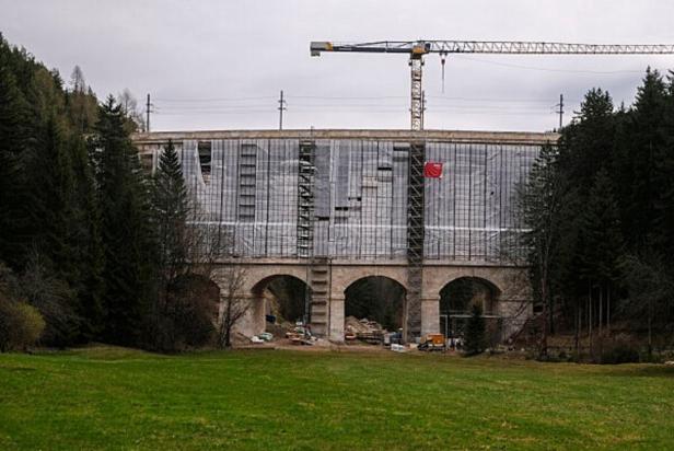 Eine Eisenbahnbrücke wird von einem Baugerüst und Planen umgeben, darüber steht ein großer Baukran, umgeben von Wald.