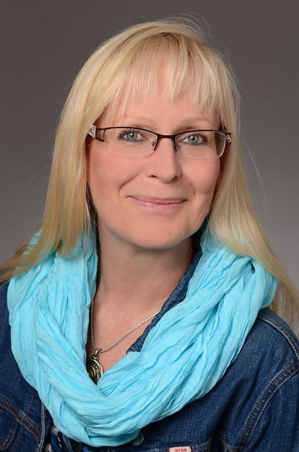 Frau mit langen blonden Haaren, Brille, hellblauem Schal und Jeansjacke vor grauem Hintergrund.