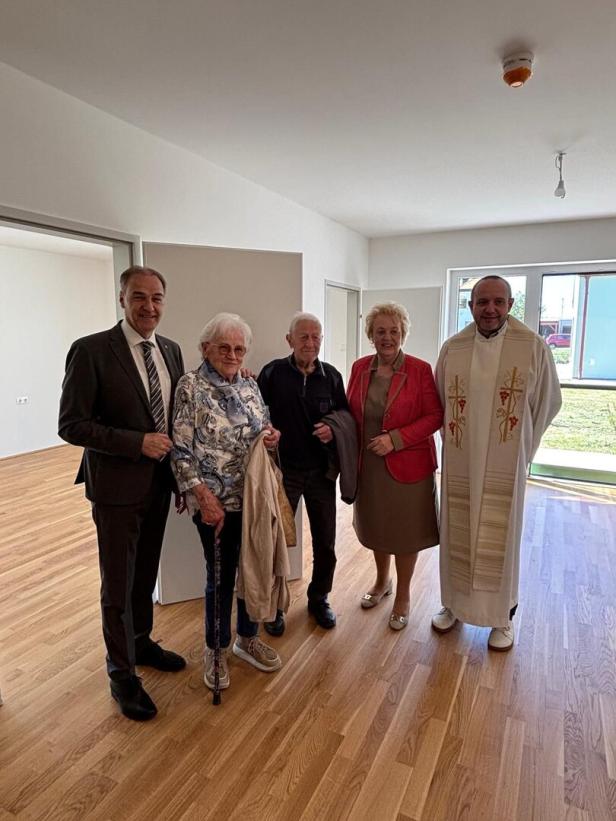 Fünf Personen stehen lächelnd in einem hellen Raum mit Holzfußboden, darunter ein Priester in liturgischer Kleidung.