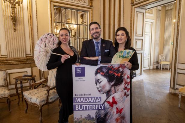 Drei Personen präsentieren im prunkvollen Raum ein Plakat zu Puccinis „Madama Butterfly“.