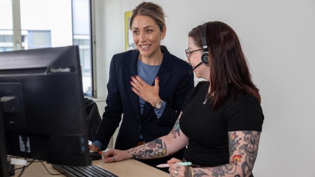 Eine blonde Frau steht mit einer schwarzhaarigen Frau mit Headset an einem PC 