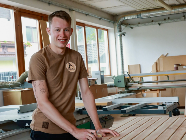 Junger Mann in einer modernen Tischlerei arbeitet an einer Holzplatte und lächelt in die Kamera.