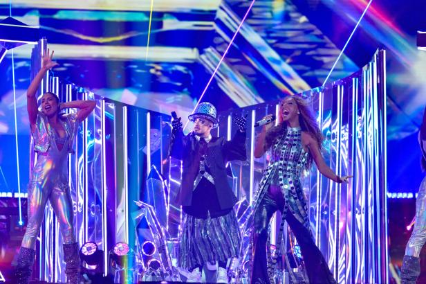 Drei Personen in glitzernden Outfits performen auf einer bunt beleuchteten Bühne mit futuristischen Lichteffekten.