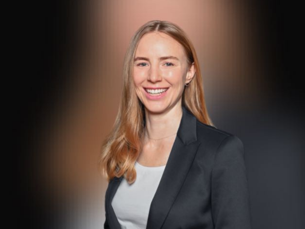 Junge Frau mit langen blonden Haaren im Business-Outfit lächelt vor dunklem Hintergrund.