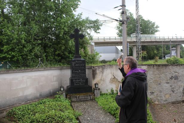 Ein Mann mit violettem Schal segnet ein Grabmal mit Kreuz auf einem kleinen Friedhof neben einer Mauer.