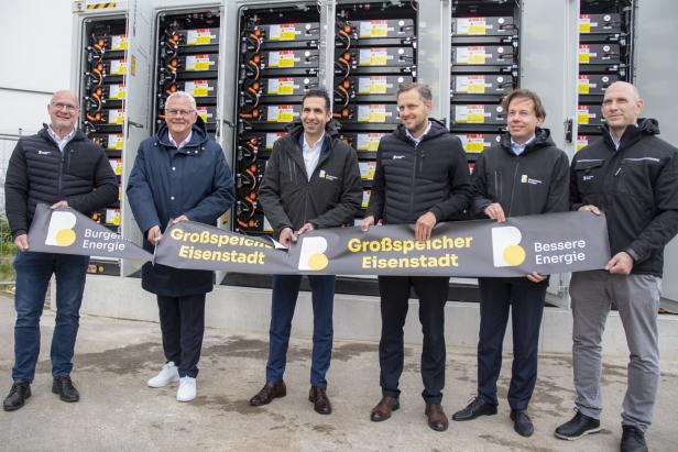 Sechs Männer stehen vor geöffneten Batterieschränken und halten gemeinsam ein Banner mit der Aufschrift „Großspeicher Eisenstadt“.