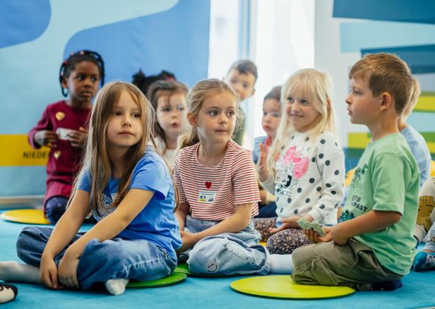 Mehrere Kinder sitzen aufmerksam auf bunten Sitzkissen in einer Bibliothek.