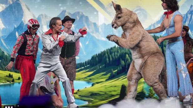 Mehrere Menschen in bunten Kostümen stehen vor einer Alpenlandschaft, einer davon kämpft spielerisch mit einem Känguru im Kostüm.