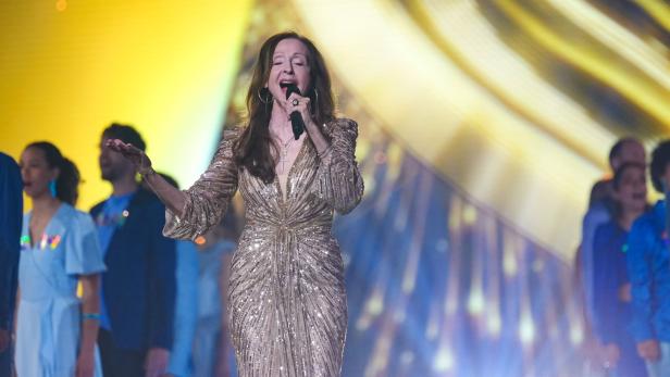 Eine Frau im glitzernden goldenen Kleid singt auf einer Bühne, im Hintergrund stehen mehrere Menschen.