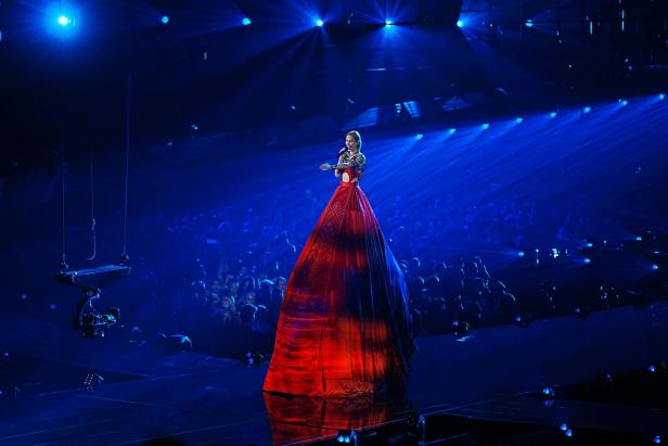 Eine Sängerin steht auf einer Bühne in einem langen, roten Kleid, umgeben von blauem Licht und Publikum.