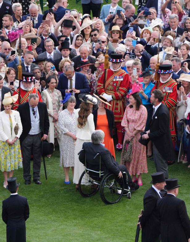 Eine Menschenmenge, darunter Prinzessin Kate und einige Leute in traditionellen Uniformen, versammelt sich auf einer grünen Rasenfläche.