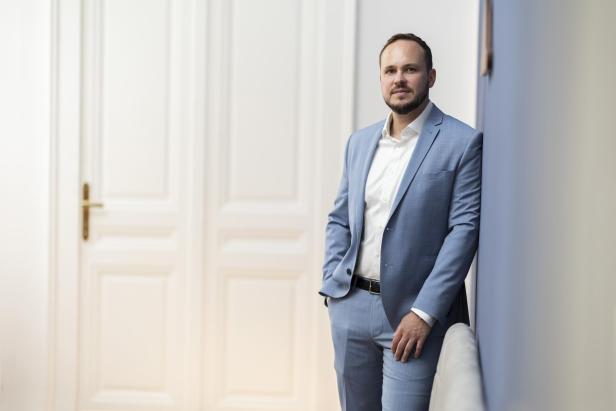 Ein Mann im hellblauen Anzug lehnt an einer Wand vor einer weißen Tür.