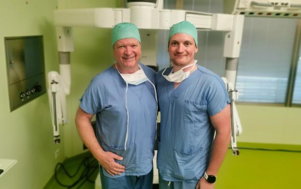 Zwei Chirurgen in OP-Kleidung stehen vor einem robotischen Assistenzsystem im Operationssaal.
