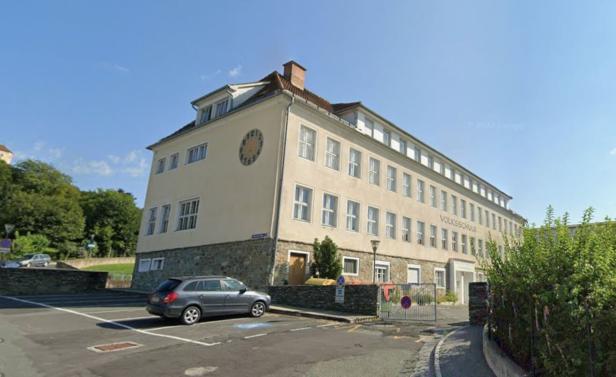 Ein langes, helles Schulgebäude mit vielen Fenstern, einer großen Uhr an der Ecke und dem Schriftzug „Volksschule“ an der Fassade.