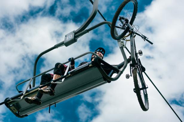 Eine Person mit Fahrradhelm sitzt in einem Sessellift, neben ihr hängt ein Mountainbike, der Himmel ist bewölkt.