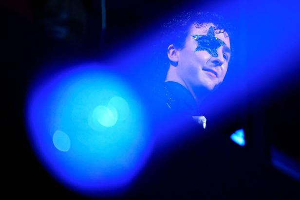 Eine Person mit glitzerndem Stern im Gesicht steht im blauen Licht und schaut leicht lächelnd zur Seite.