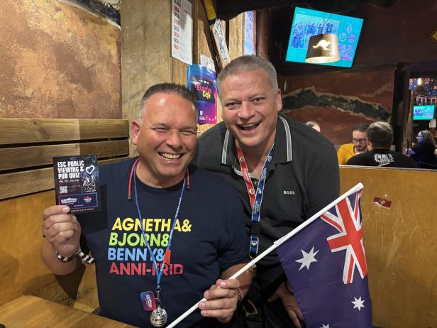 Zwei Männer lächeln mit einer australischen Flagge und einem ESC-Quiz-Flyer in einem Pub.