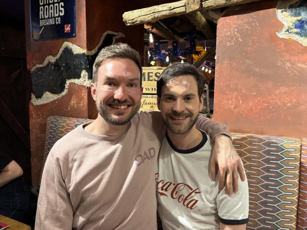 Zwei lächelnde Männer posieren, einer trägt ein "DAD"-Sweatshirt, der andere ein Coca-Cola-T-Shirt.