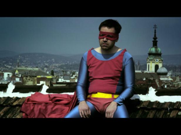 Ein Mann im Superheldenkostüm sitzt entmutigt auf einem Dach mit Blick auf die Stadt.