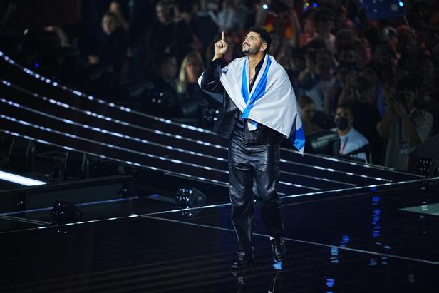 Ein Mann mit einer blau-weißen Flagge um die Schultern steht auf einer Bühne und zeigt mit dem Finger nach oben.
