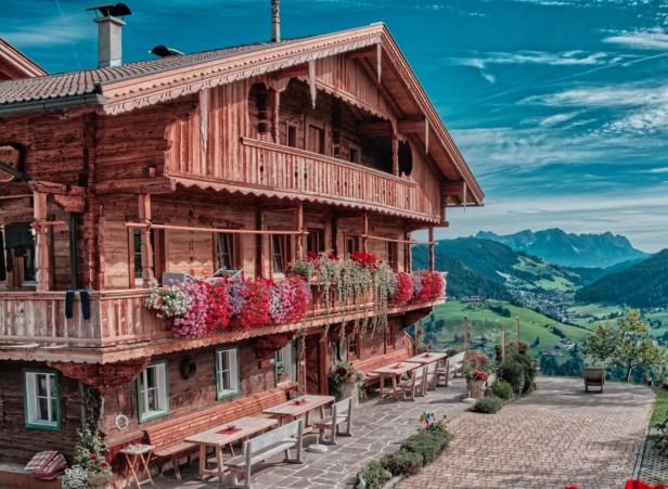 Traditionelles Holzhaus mit bunten Blumen am Balkon, Sitzbänken davor und Blick auf grüne Berge und ein Tal.