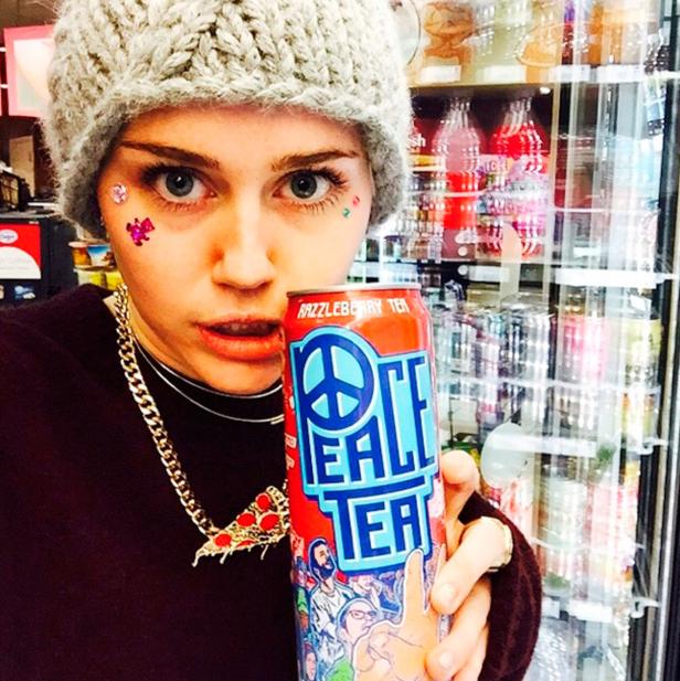 Eine Person mit Mütze und Gesichtsschmuck hält eine Dose „Peace Tea“ vor einem Kühlschrank.