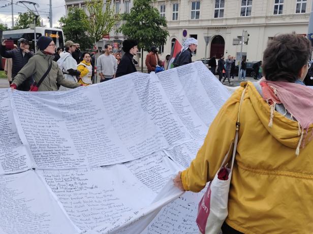 Mehrere Menschen halten ein großes Tuch mit zahlreichen handgeschriebenen Namen, während andere im Hintergrund stehen.