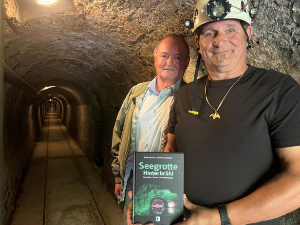 Zwei Männer stehen in einem beleuchteten Tunnel, einer trägt einen Helm und hält ein Buch mit dem Titel „Seegrotte Hinterbrühl“.