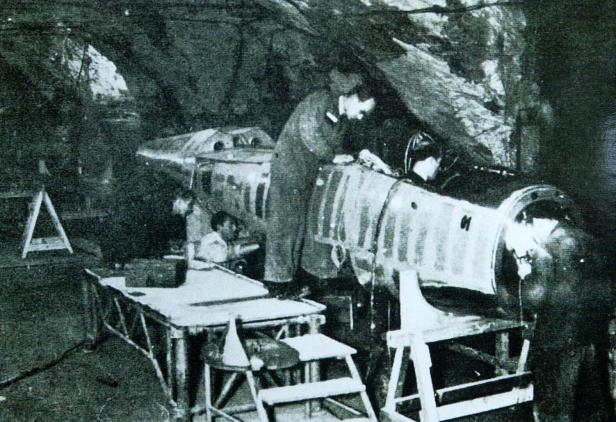 Mehrere Personen arbeiten in einer unterirdischen Werkstatt an einem großen metallischen Flugzeugrumpf.