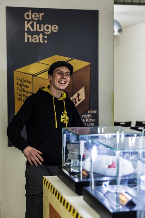 Ein junger Mann mit schwarzem Hoodie und Mütze steht lachend neben einer Vitrine vor einem Poster mit Vorratsliste.