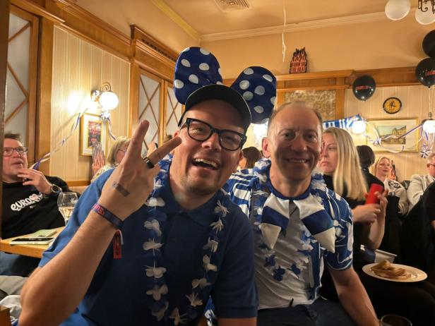 Zwei Männer in blau-weißer Verkleidung feiern fröhlich in einer festlich geschmückten Gruppe.
