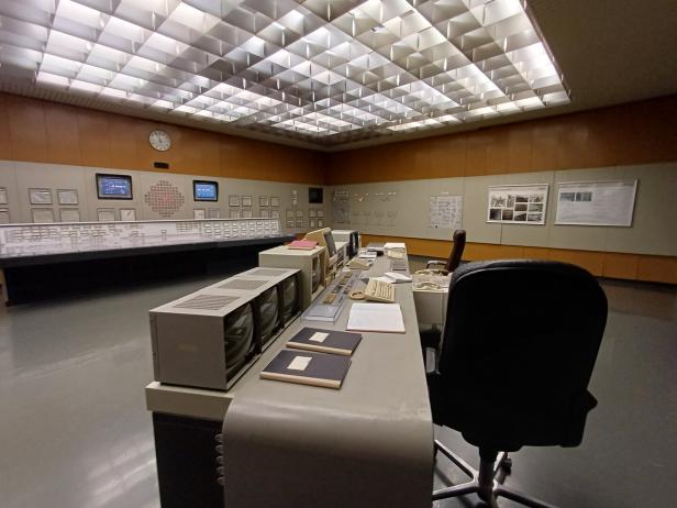 Großer Kontrollraum mit Schreibtischen, Monitoren, Telefonen, Tastaturen und vielen Schaltern an den Wänden.