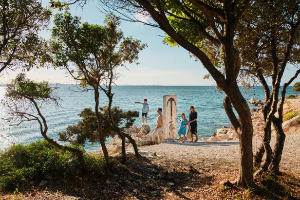 Familie spaziert an einem sonnigen Tag am Meer entlang, umgeben von Bäumen und Felsen.