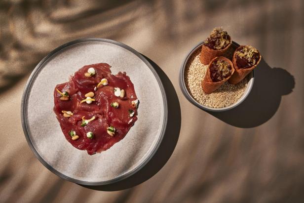 Fein angerichtetes Thunfisch-Carpaccio und drei gefüllte Waffelröllchen auf hellem Untergrund.