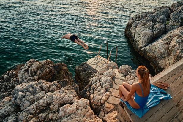 Mann springt von Felsen ins Meer, Frau sitzt entspannt auf einem Handtuch am Ufer.