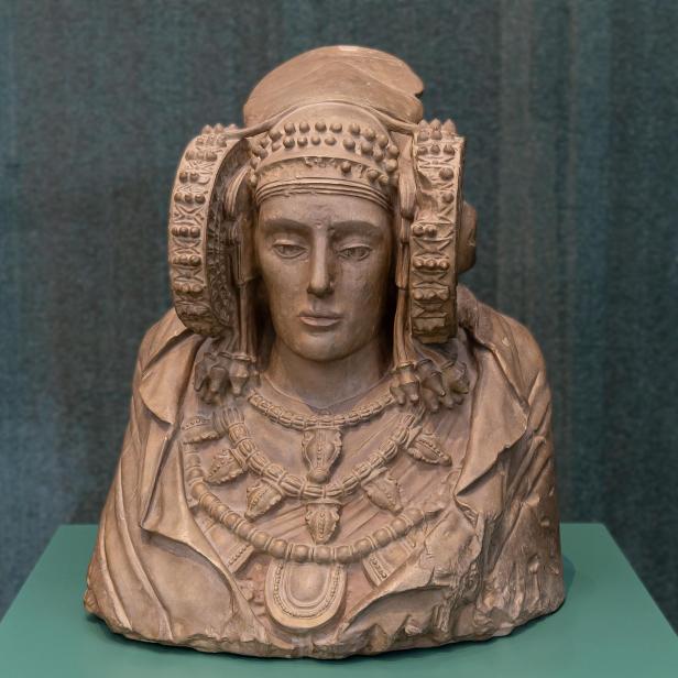 Büste der Dama de Elche mit aufwendigem Kopfschmuck und Schmuck auf grünem Sockel ausgestellt.