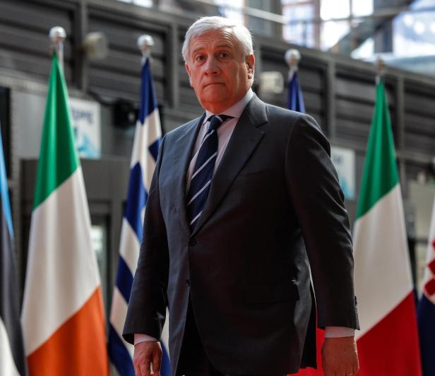 Ein älterer Mann im Anzug steht vor mehreren europäischen Nationalflaggen in einem modernen Gebäude.