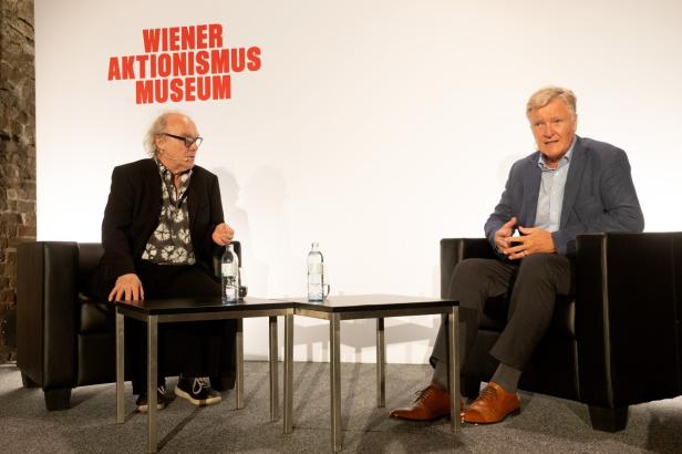 Zwei Personen sitzen auf schwarzen Sesseln mit einem kleinen schwarzen Tisch und Wasserflaschen dazwischen vor einer Wand mit roter Aufschrift 'WIENER AKTIONISMUS MUSEUM'.