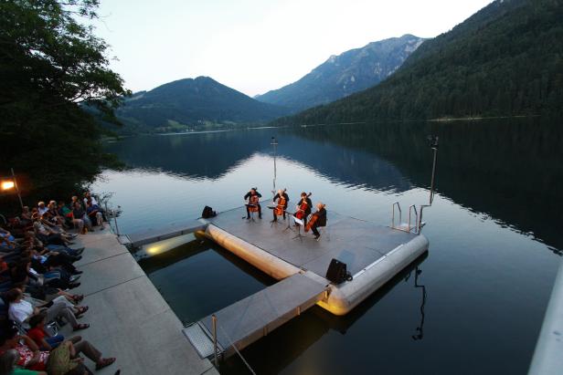 Vier Musiker spielen auf einem schwimmenden Podest auf einem See vor Zuschauern, umgeben von Bergen.