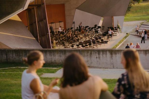 Drei Personen sitzen auf einer Wiese vor einer modernen Open-Air-Bühne mit Orchesterbestuhlung.